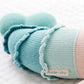 Close-up of Skye the Mermaid, a light blue knitted stuffed animal by Cuddle + Kind, featuring its label and lying on a white textured surface.