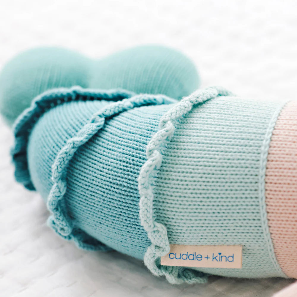 Close-up of Skye the Mermaid, a light blue knitted stuffed animal by Cuddle + Kind, featuring its label and lying on a white textured surface.
