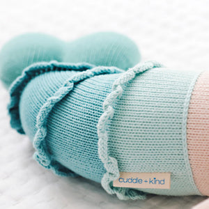 Close-up of Skye the Mermaid, a light blue knitted stuffed animal by Cuddle + Kind, featuring its label and lying on a white textured surface.