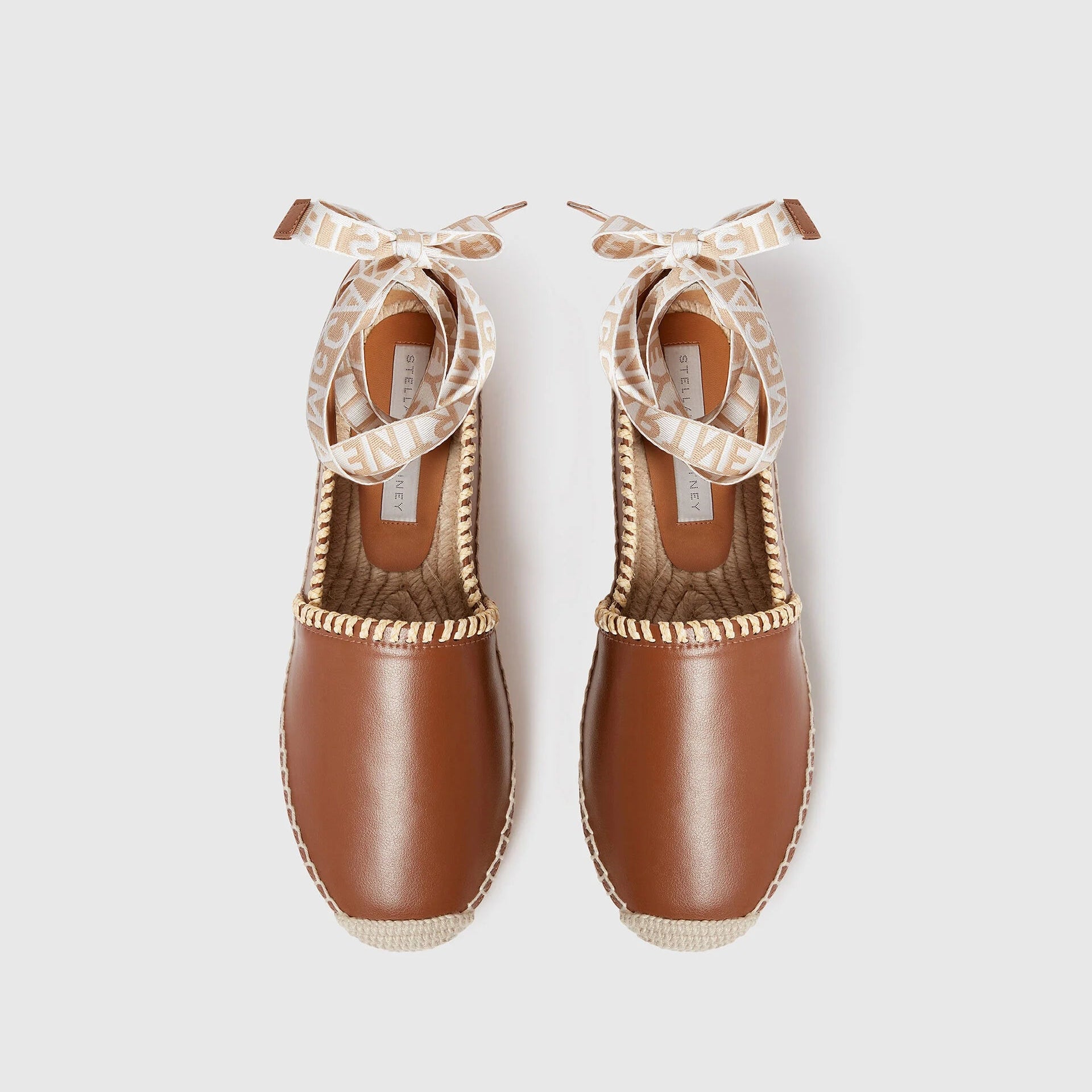 The Gaia Whipstitched Flat by Stella McCartney is a pair of brown leather espadrilles with woven soles and beige ribbon ankle ties, showcasing natural textures and a handcrafted finish, displayed side by side on a white background.