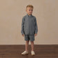 A young boy wearing the Rylee & Cru Boys’ Oceanside Tech Short—a gray zip-up jacket paired with matching elastic waistband shorts featuring a convenient zip back pocket—stands on a wooden floor against a beige background, looking slightly to his right and highlighting these perfect everyday essential shorts.