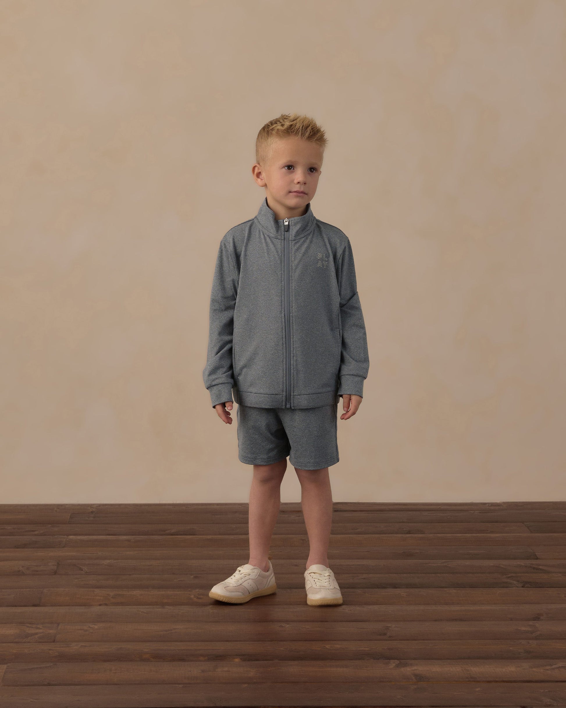 A young boy wearing the Rylee & Cru Boys’ Oceanside Tech Short—a gray zip-up jacket paired with matching elastic waistband shorts featuring a convenient zip back pocket—stands on a wooden floor against a beige background, looking slightly to his right and highlighting these perfect everyday essential shorts.