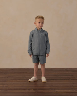 A young boy wearing the Rylee & Cru Boys’ Oceanside Tech Short—a gray zip-up jacket paired with matching elastic waistband shorts featuring a convenient zip back pocket—stands on a wooden floor against a beige background, looking slightly to his right and highlighting these perfect everyday essential shorts.