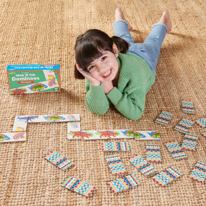 A young girl smiles at the camera while lying on a carpet, next to Eeboo’s Dinosaur Head to Toe Dominoes game spread out beside her.