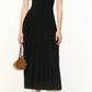 Person wearing a sleeveless black pleated Elyse Dress by Staud with a crew neckline, cream slouchy knee-high boots, and holding a small brown round handbag, standing against a plain background.