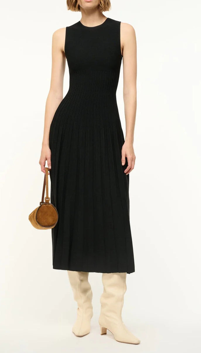 Person wearing a sleeveless black pleated Elyse Dress by Staud with a crew neckline, cream slouchy knee-high boots, and holding a small brown round handbag, standing against a plain background.