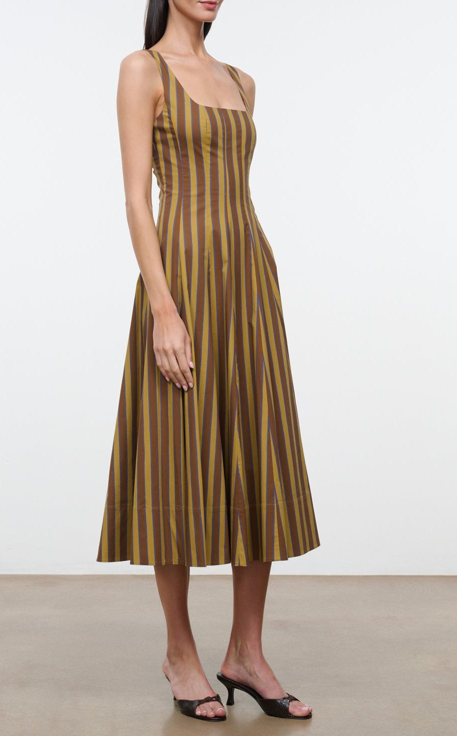 A woman stands against a plain background in heeled sandals, wearing the Staud Wells Midi Cotton Poplin Dress—a sleeveless, calf-length style featuring vertical brown and yellow stripes that showcase the effortlessly polished signature silhouette of Staud.