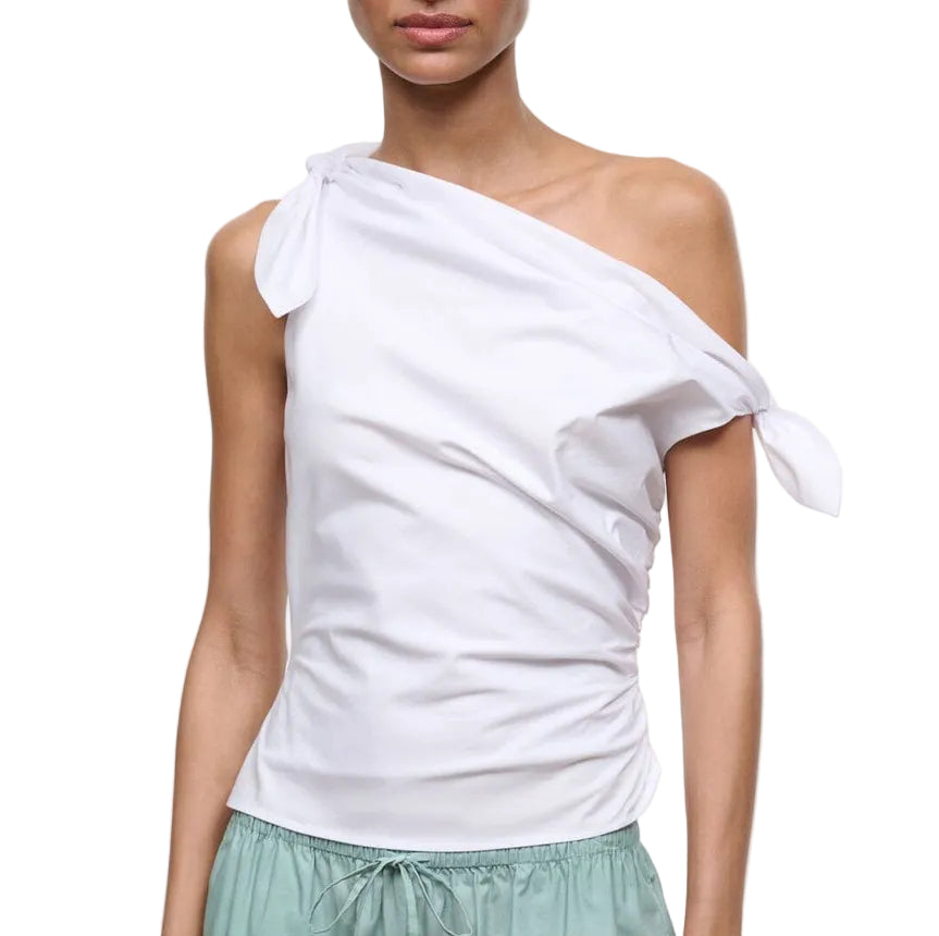 A person wearing the Staud Bellaria Top—an elegant white evening piece with an asymmetrical neckline and tied knots at the shoulder and sleeve—paired with light green pants.