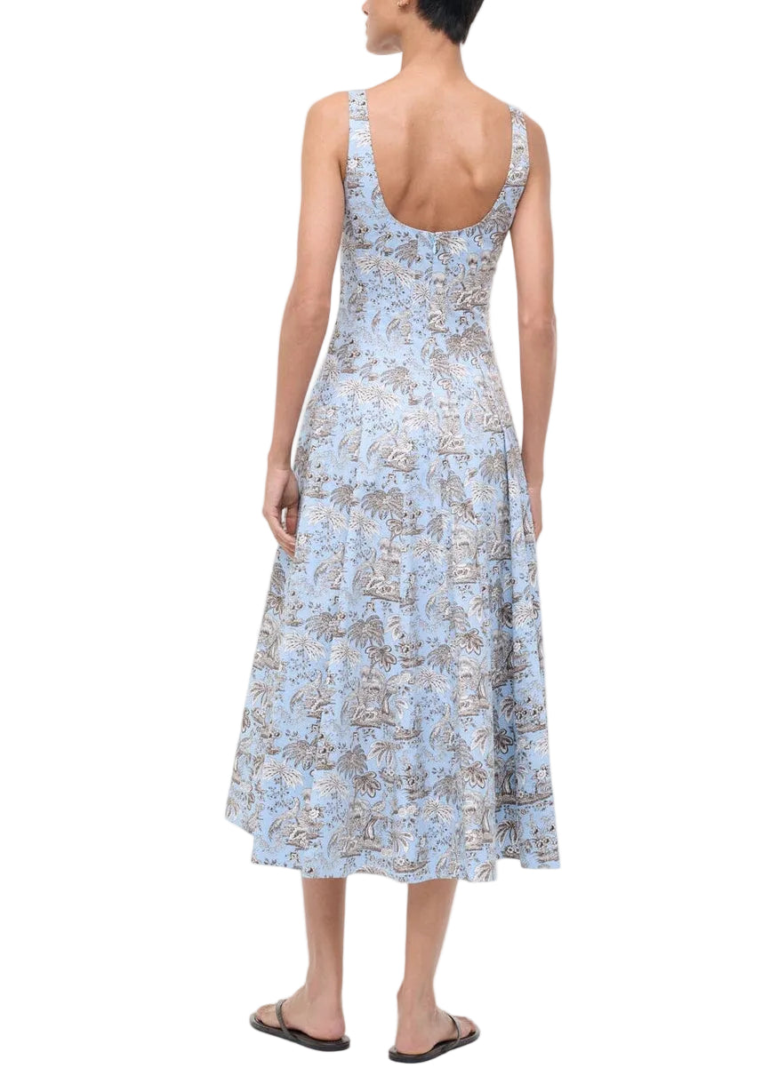 A person wears the Staud Wells Dress Sky Toile, a sleeveless light blue printed cotton poplin dress, with black flip-flops, seen from the back against a plain white background.