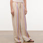 A person wears the Staud Mateo Linen Pant—relaxed fit, beige with vertical multicolored stripes—paired with a white tank top and black sandals, standing on a neutral floor against a plain background.