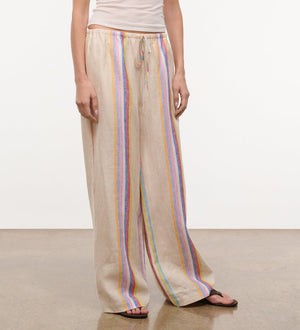 A person wears the Staud Mateo Linen Pant—relaxed fit, beige with vertical multicolored stripes—paired with a white tank top and black sandals, standing on a neutral floor against a plain background.