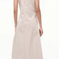 A person faces away, wearing the Staud Viola Dress with a draped skirt and ankle-length beige fabric, paired with low-heeled snakeskin shoes against a plain background.