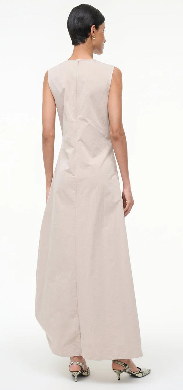 A person faces away, wearing the Staud Viola Dress with a draped skirt and ankle-length beige fabric, paired with low-heeled snakeskin shoes against a plain background.