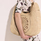 A person in a white dress with a cherry pattern holds the Stella McCartney Logo Raffia Slouchy Large Tote Bag, which features circular logo lettering on the front.
