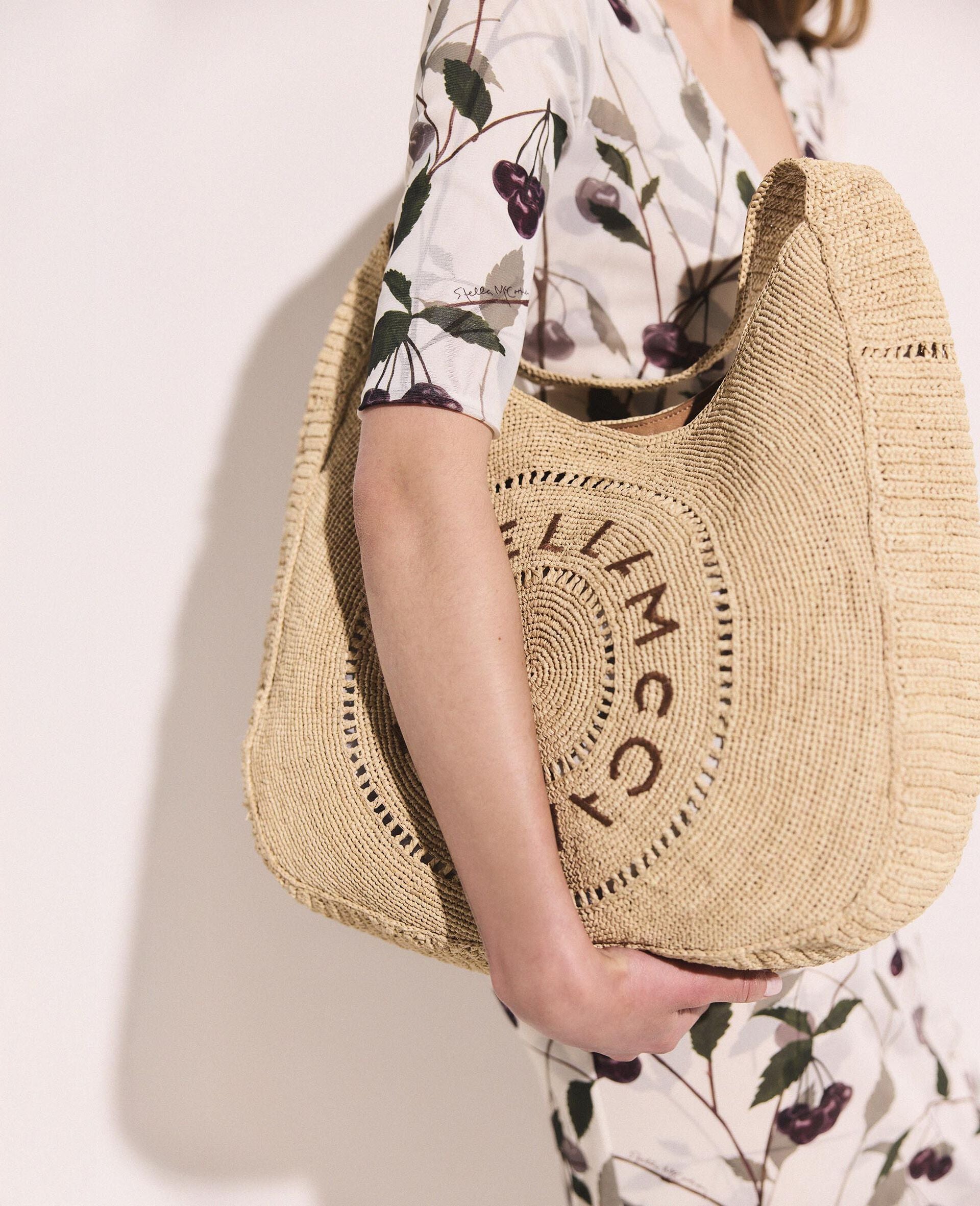 A person in a white dress with a cherry pattern holds the Stella McCartney Logo Raffia Slouchy Large Tote Bag, which features circular logo lettering on the front.