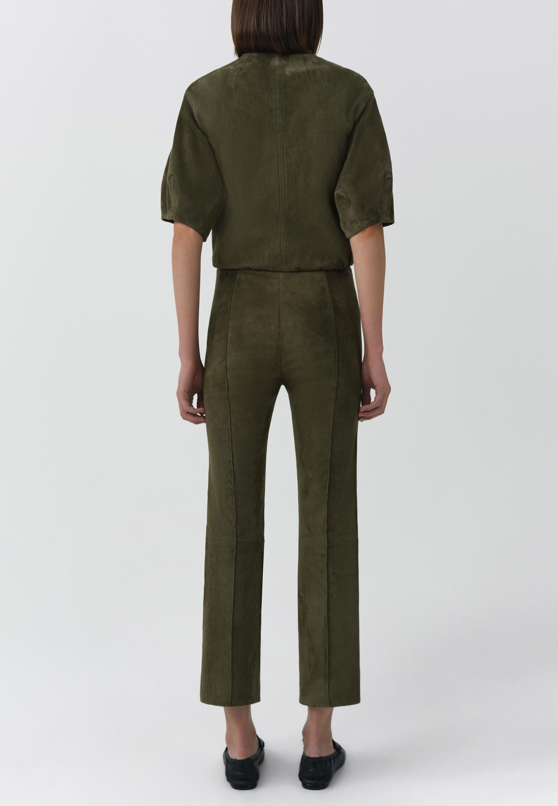 A person stands facing away, wearing the Simkhai Austyn Short Sleeve Suede Jacket in olive green with matching pants and black shoes, set against a plain light background.