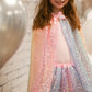 A young girl wearing the Great Pretenders Ombre Sequin Cape and matching tutu smiles in front of a white brick wall with silver balloons, showing off her favorite accessory.