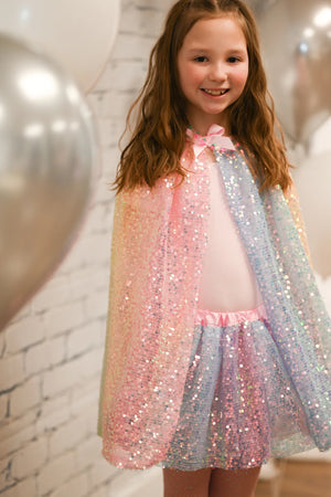 A young girl wearing the Great Pretenders Ombre Sequin Cape and matching tutu smiles in front of a white brick wall with silver balloons, showing off her favorite accessory.