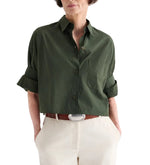 A person wears a TWP Next Ex Superfine Cotton Shirt in green, cropped short-sleeve and button-down style, tucked into high-waisted light beige pants with a brown belt featuring a silver buckle.