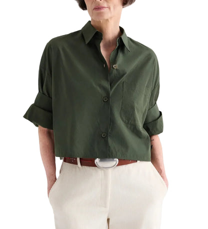 A person wears a TWP Next Ex Superfine Cotton Shirt in green, cropped short-sleeve and button-down style, tucked into high-waisted light beige pants with a brown belt featuring a silver buckle.