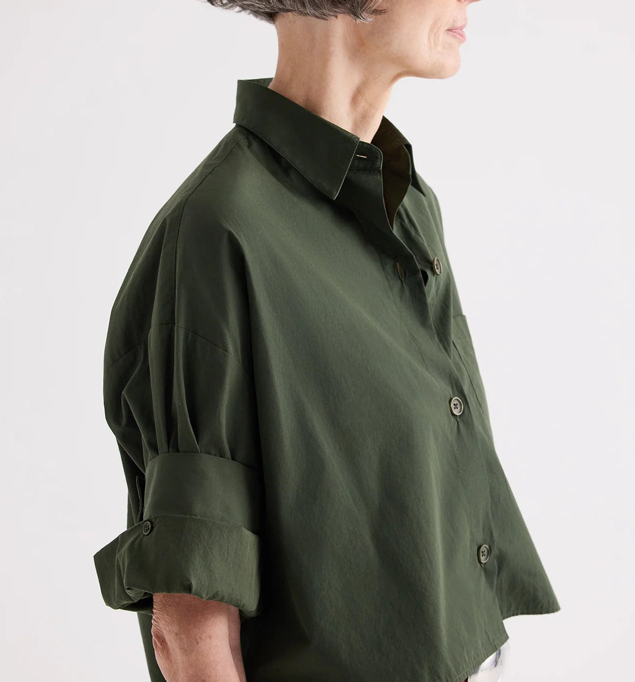 A person with short gray hair wears the TWP Next Ex Superfine Cotton Shirt in Rosen Green, a loose-fitting, cropped, short-sleeve button-down with rolled-up sleeves, shown from the side against a plain background.
