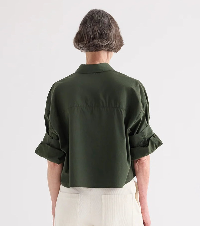 A person with short gray hair faces away from the camera, wearing the TWP Next Ex Superfine Cotton Shirt in Rosen Green and light-colored pants.