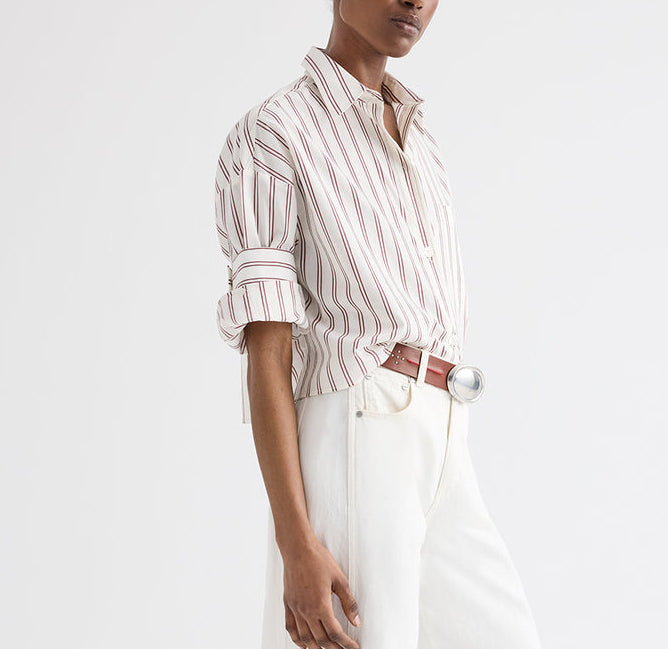A person wearing the TWP Next Ex Stripe Shirt, a white and red vertical striped cotton-silk cropped short-sleeve button-down with rolled sleeves, tucked into high-waisted white pants and accessorized with a brown belt featuring a large silver buckle.
