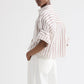 A person stands sideways with hands in pockets against a plain background, wearing TWP's Next Ex Stripe Shirt—a cropped, short-sleeve button-down—paired with white pants.