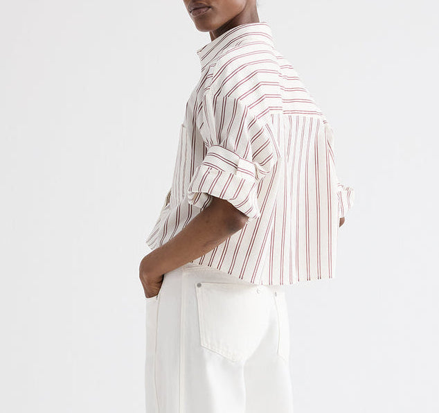 A person stands sideways with hands in pockets against a plain background, wearing TWP's Next Ex Stripe Shirt—a cropped, short-sleeve button-down—paired with white pants.