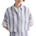 A woman wearing the TWP Next Ex Linen Stripe Shirt—a loose, ivory button-up shirt with vertical navy and white stripes, rolled-up sleeves, and a front pocket—paired with matching striped pants.