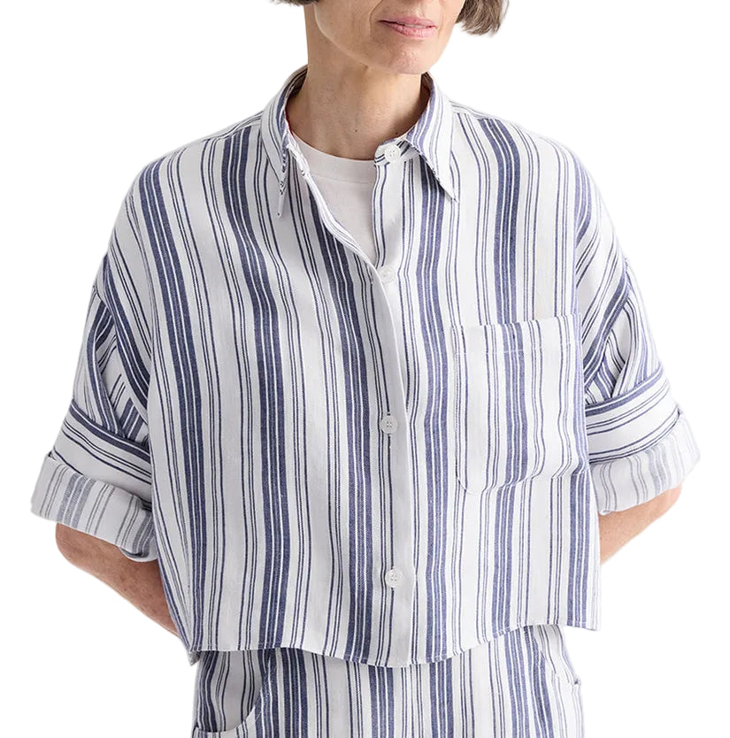 A woman wearing the TWP Next Ex Linen Stripe Shirt—a loose, ivory button-up shirt with vertical navy and white stripes, rolled-up sleeves, and a front pocket—paired with matching striped pants.