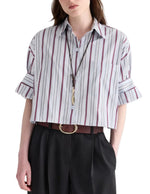 A woman wearing the TWP Next Ex Striped Superfine Poplin Shirt—a cropped, short-sleeve button-down with a men’s stripe pattern and rolled sleeves—paired with black trousers, a brown belt, and a long necklace.