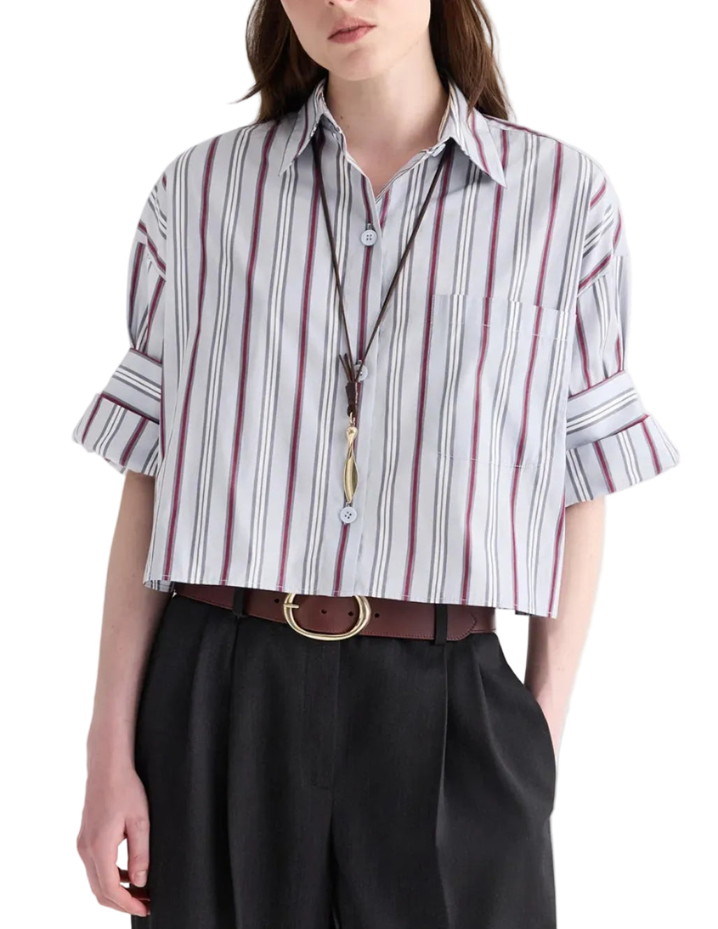 A woman wearing the TWP Next Ex Striped Superfine Poplin Shirt—a cropped, short-sleeve button-down with a men’s stripe pattern and rolled sleeves—paired with black trousers, a brown belt, and a long necklace.