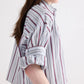 A person stands sideways against a plain light background, wearing the TWP Next Ex Striped Superfine Poplin Shirt—an airy, long-sleeve white shirt featuring vertical maroon and gray men’s stripes, with the sleeves casually rolled up.