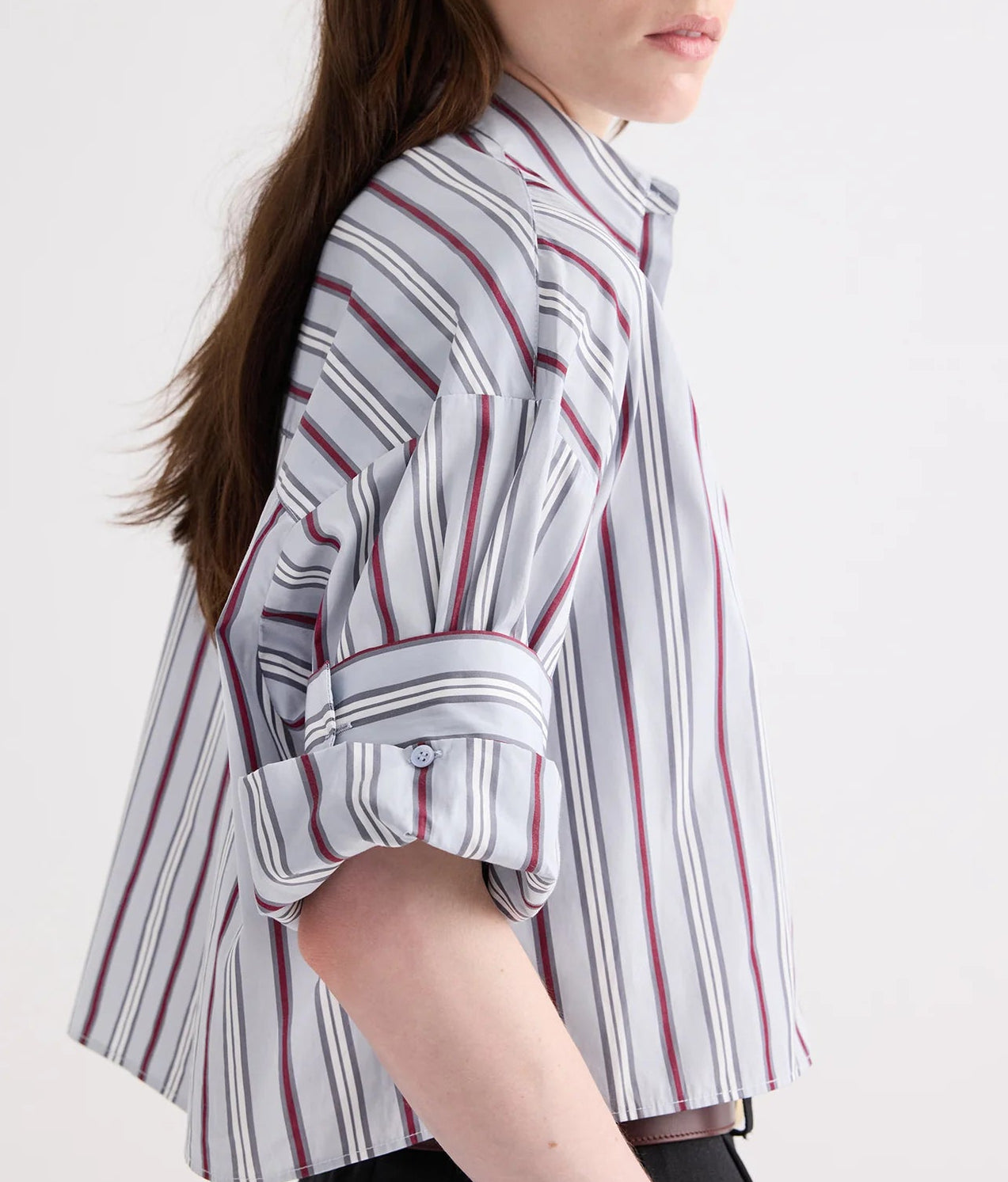 A person stands sideways against a plain light background, wearing the TWP Next Ex Striped Superfine Poplin Shirt—an airy, long-sleeve white shirt featuring vertical maroon and gray men’s stripes, with the sleeves casually rolled up.