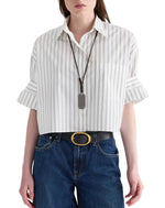 A person is wearing the TWP Next Ex Mixed Hairline Stripe Shirt—a white, lightweight cotton, cropped short-sleeve button-down with a subtle hairline stripe—tucked into blue jeans with a black belt featuring a gold buckle, and accessorized with a long necklace.