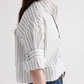 A person is shown from the side against a plain background, wearing the TWP Next Ex Mixed Hairline Stripe Shirt—a lightweight cotton cropped short-sleeve button-down featuring hairline stripes and rolled sleeves—paired with blue jeans.
