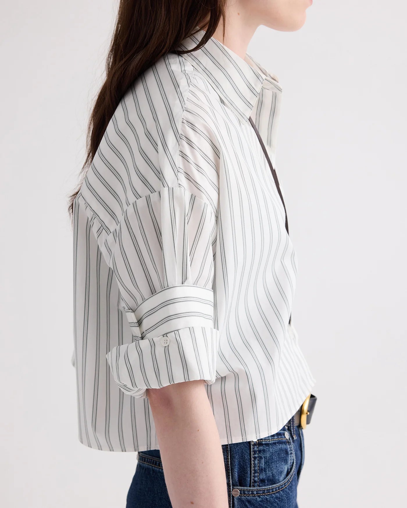 A person is shown from the side against a plain background, wearing the TWP Next Ex Mixed Hairline Stripe Shirt—a lightweight cotton cropped short-sleeve button-down featuring hairline stripes and rolled sleeves—paired with blue jeans.