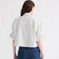 A person with long brown hair, wearing the TWP Next Ex Mixed Hairline Stripe Shirt—a lightweight cotton white button-up shirt featuring hairline vertical stripes and rolled sleeves—paired with blue jeans, stands facing away from the camera against a plain background.