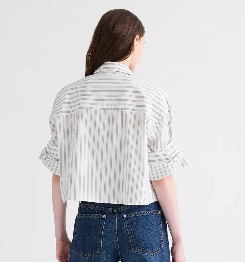 A person with long brown hair, wearing the TWP Next Ex Mixed Hairline Stripe Shirt—a lightweight cotton white button-up shirt featuring hairline vertical stripes and rolled sleeves—paired with blue jeans, stands facing away from the camera against a plain background.