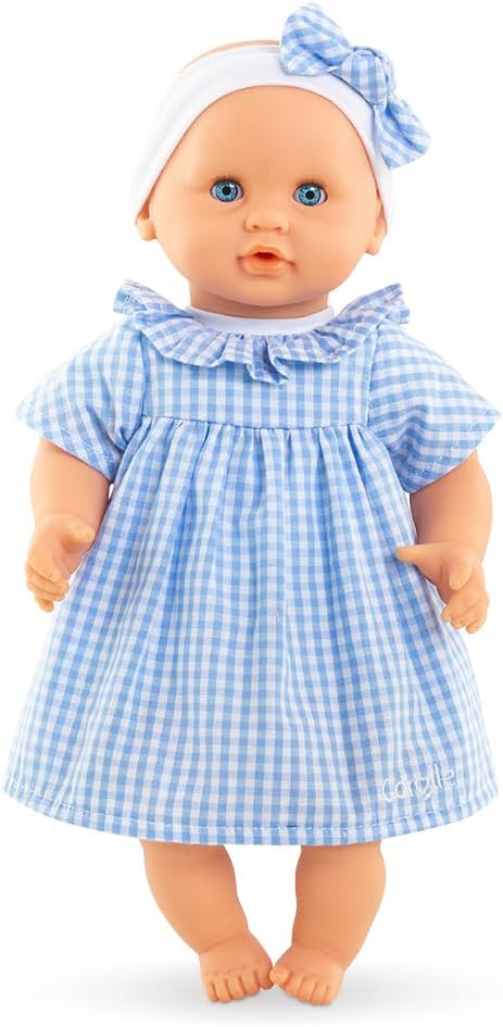 The Corolle Premier Calin - Milly Doll is a soft-bodied baby doll dressed in a blue and white gingham dress with a matching headband and bow, shown standing upright against a white background.