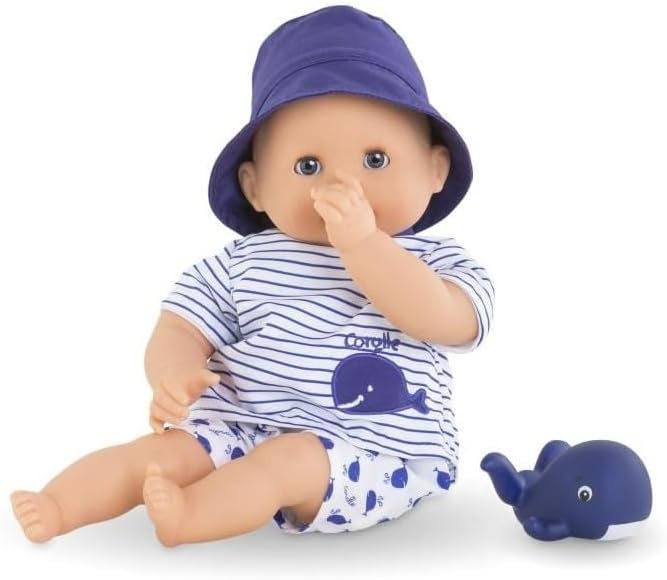 The Corolle Bebe Bath Doll, dressed in a blue hat and striped whale-print outfit, sits with one thumb in its mouth beside a blue toy whale—an ideal water-play companion.
