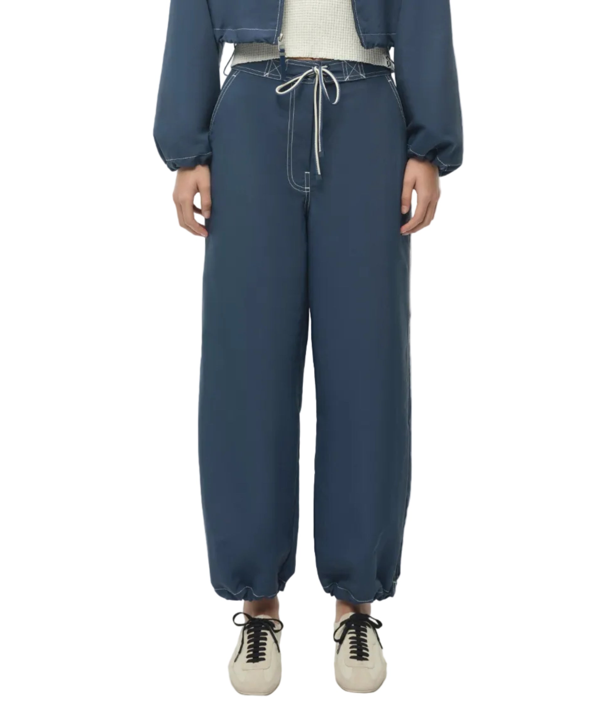 A person wears Simkhai's Lennox Cotton Straight Leg Pant in dark indigo with an adjustable drawstring waistband, styled with a matching blue jacket and white sneakers, standing against a white background.