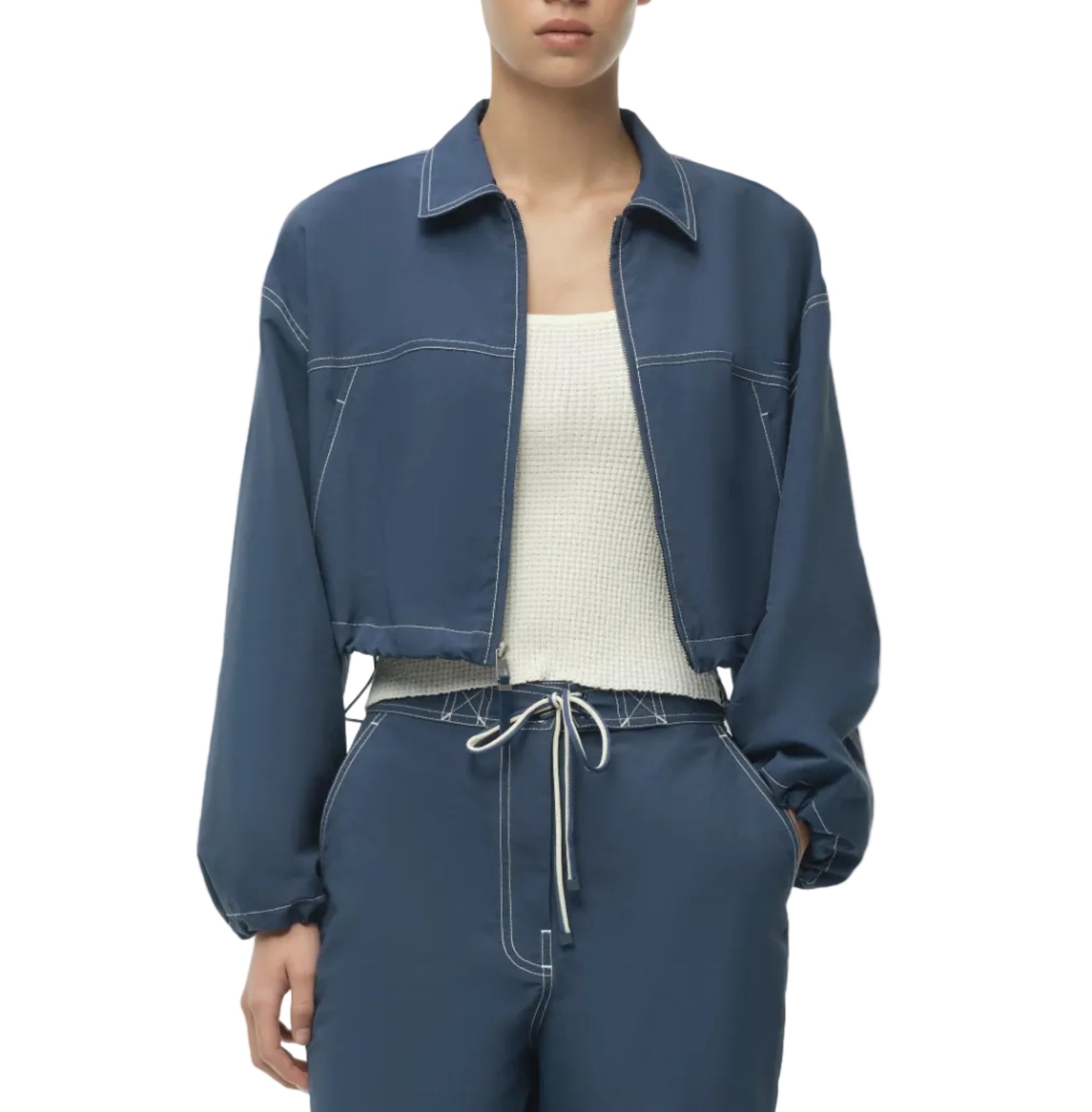 A person wearing the Simkhai Colsen Cotton Cropped Jacket in dark indigo with white stitching, paired with a white textured cotton top and blue drawstring pants.