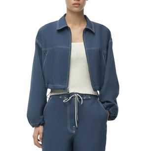 A person wearing the Simkhai Colsen Cotton Cropped Jacket in dark indigo with white stitching, paired with a white textured cotton top and blue drawstring pants.