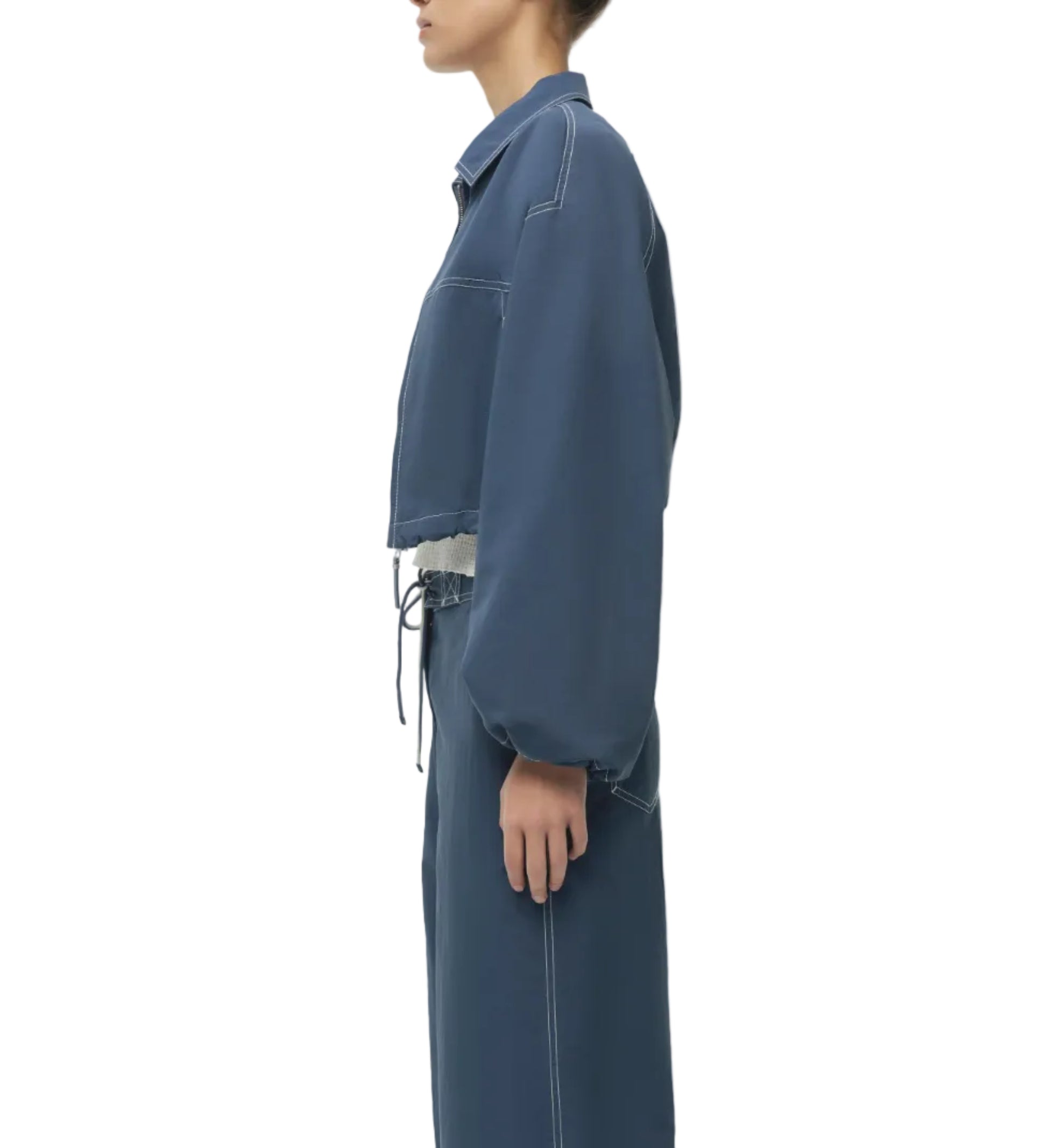 A person stands in profile wearing the Simkhai Colsen Cotton Cropped Jacket in dark indigo with white stitching, paired with matching high-waisted pants, both showcasing a boxy silhouette against a white background.