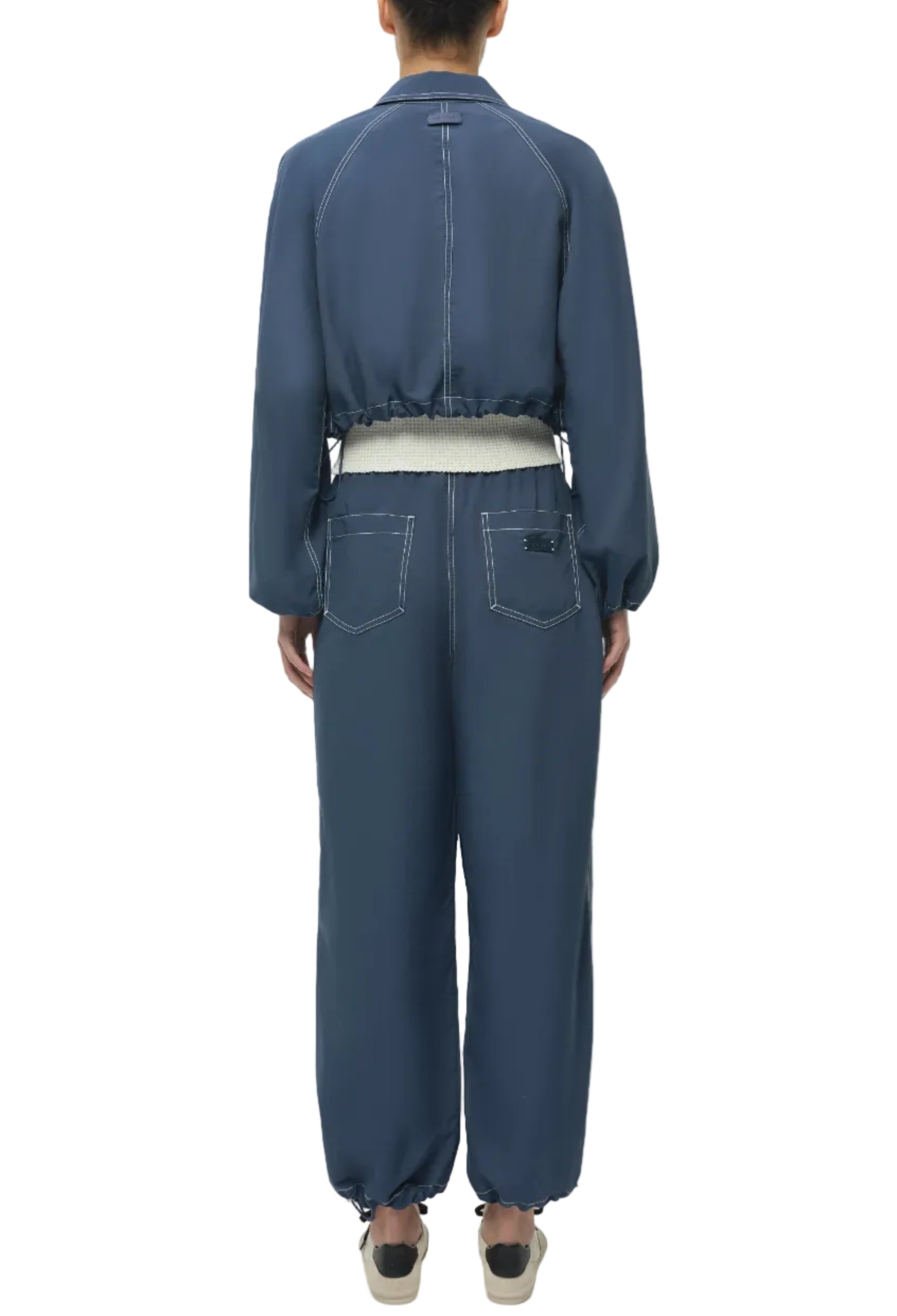 A person stands facing away, wearing the Simkhai Colsen Cotton Cropped Jacket, featuring dark indigo fabric with white contrast stitching, an elastic waistband, and cuffs, paired with slip-on shoes.