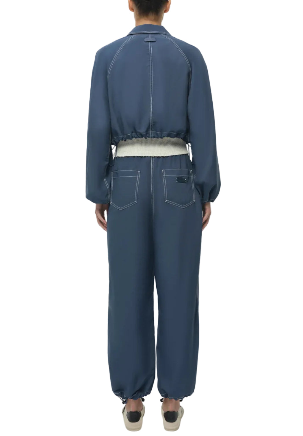 A person stands facing away, wearing Simkhai's Lennox Cotton Straight Leg Pant in Dark Indigo with white contrast stitching, elastic waistband with adjustable drawstring, cuffed ankles, and slip-on shoes.