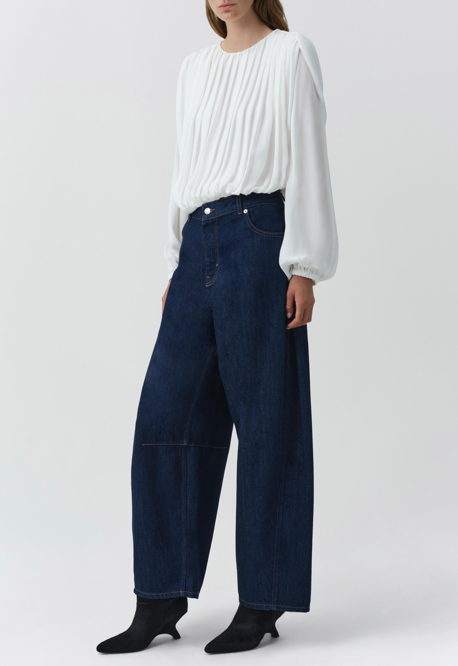 A person wears the Simkhai Frida Top, a white long-sleeve silk georgette blouse with pleats and blouson sleeves, paired with wide-leg dark blue jeans, standing against a plain light background.