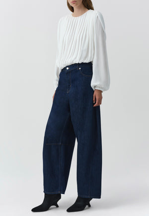 A person wears the Simkhai Frida Top, a white long-sleeve silk georgette blouse with pleats and blouson sleeves, paired with wide-leg dark blue jeans, standing against a plain light background.